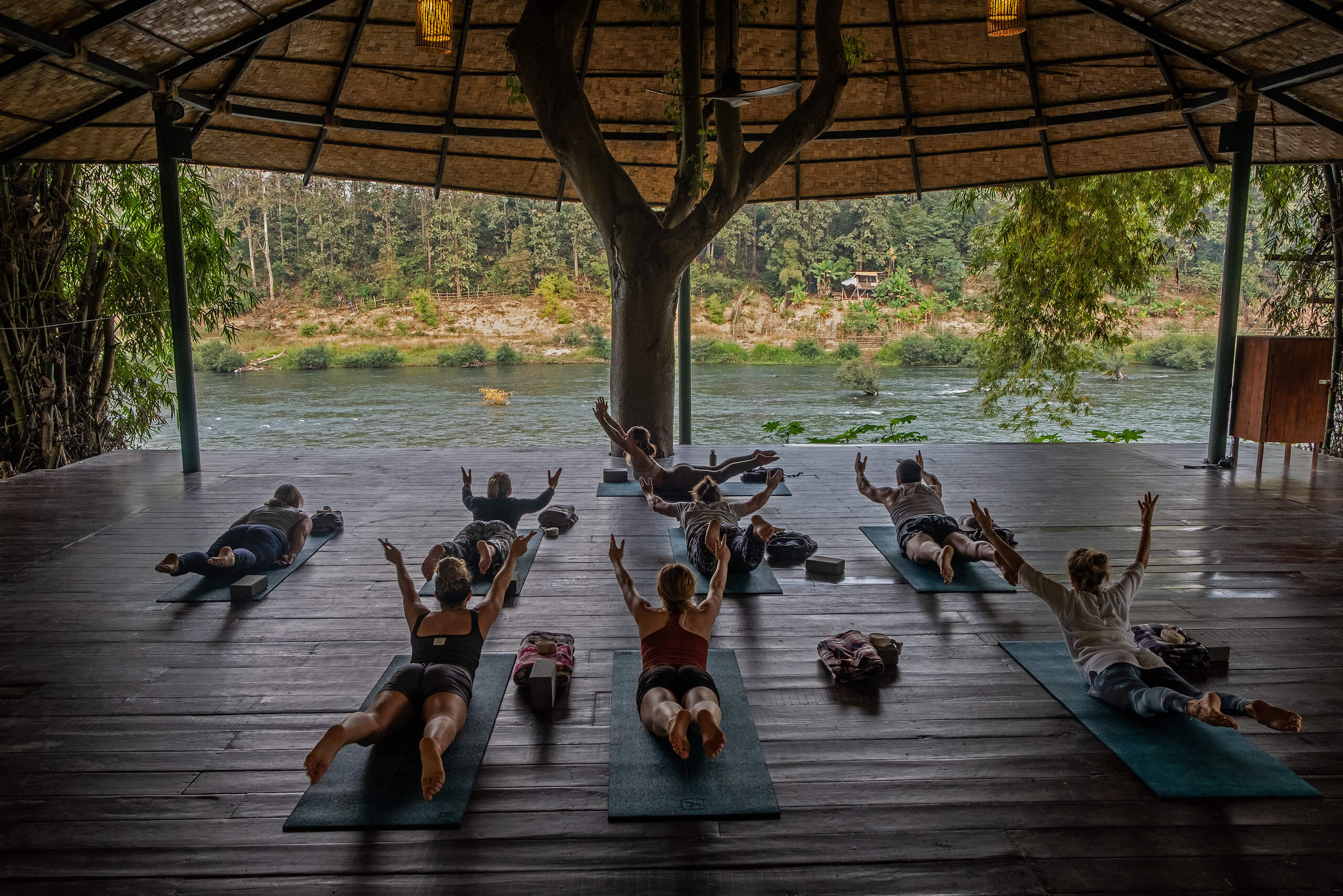 Group of people practicing yoga on mats in an open pavilion by the Namkhan river in Luang Prabang, with instructors leading the session at the front.