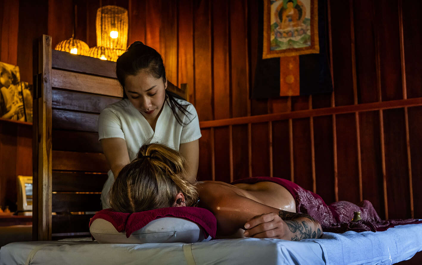 A massage therapist giving a back massage to a woman lying face down on a massage table in a dimly lit room with wooden decor, overlooking the serene Namkhan River in Luang Prabang