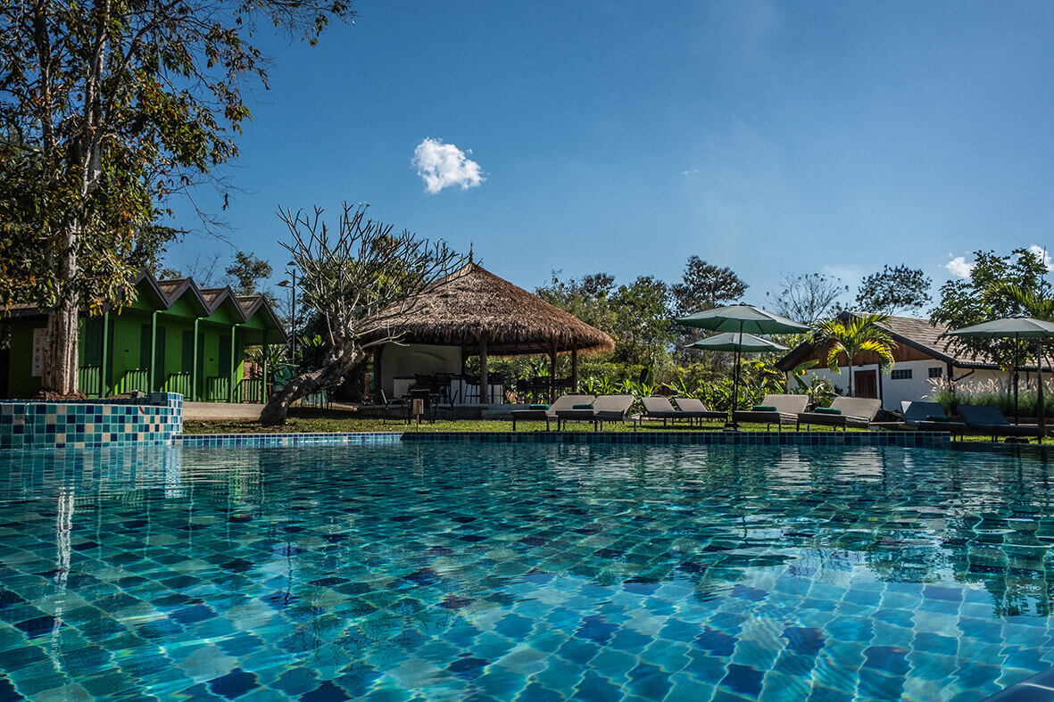 A tranquil resort pool with blue mosaic tiles, surrounded by lounge chairs, a thatched-roof hut, and lush green trees under a clear blue sky in Luang Prabang.