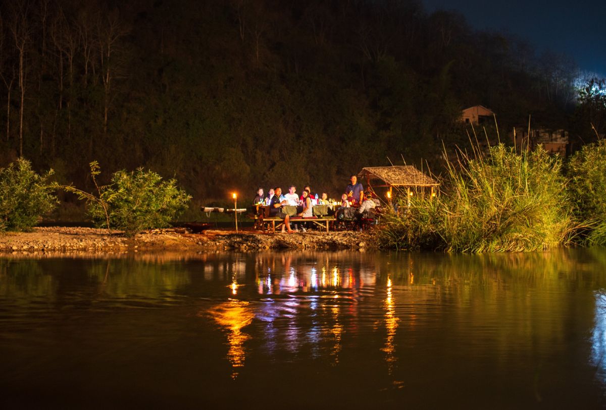A group of people enjoying a BBQ experience by a river at night.