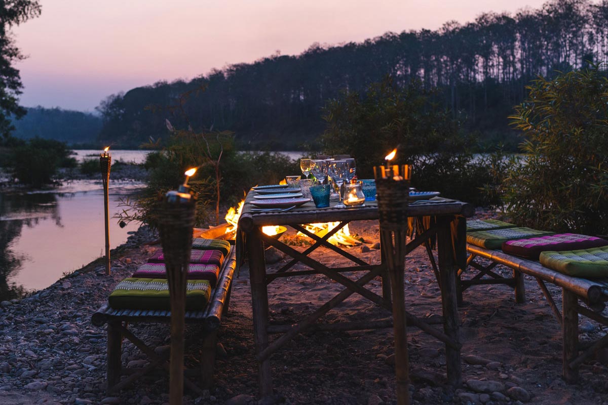 A table set up near a river at dusk, with BBQ huts nearby.