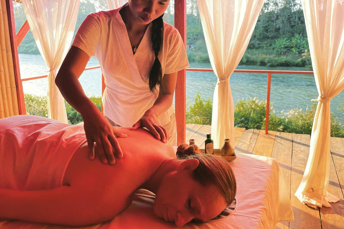 A woman experiencing wellbeing, receiving a massage on a bed in front of a tranquil river.