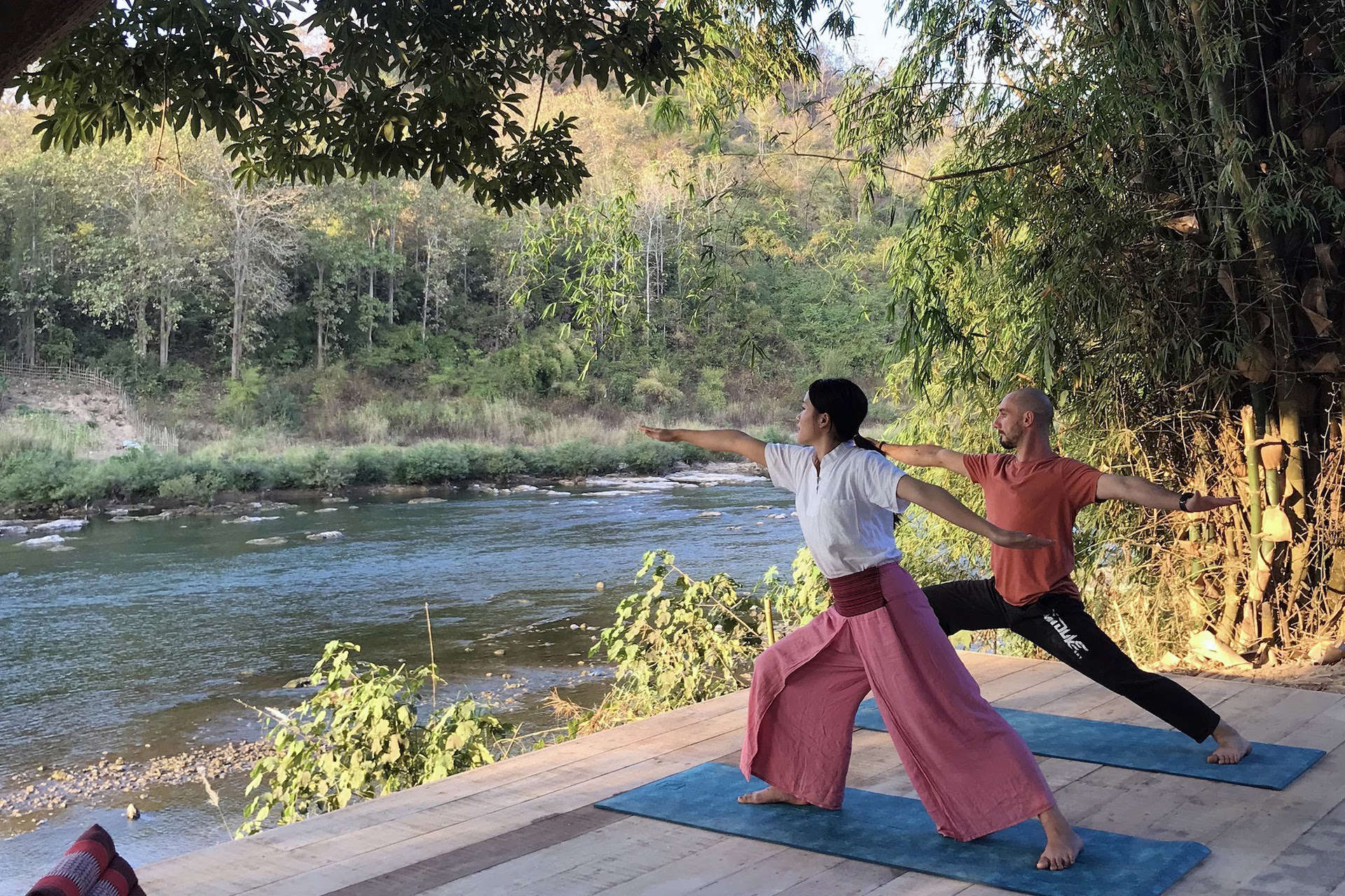 Two people practicing yoga on a wooden deck near a river in Luang Prabang.