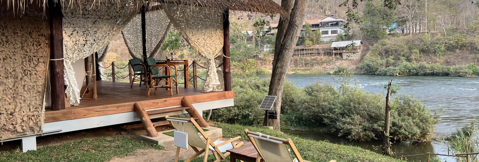 A luang prabang hut next to a river with chairs and a hammock.