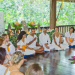 Group of people dressed in white, sitting in a circle at a yoga retreat at Namkhan Eco Farm in Luang Prabang, with flower decorations on the floor in a wooden-floored
