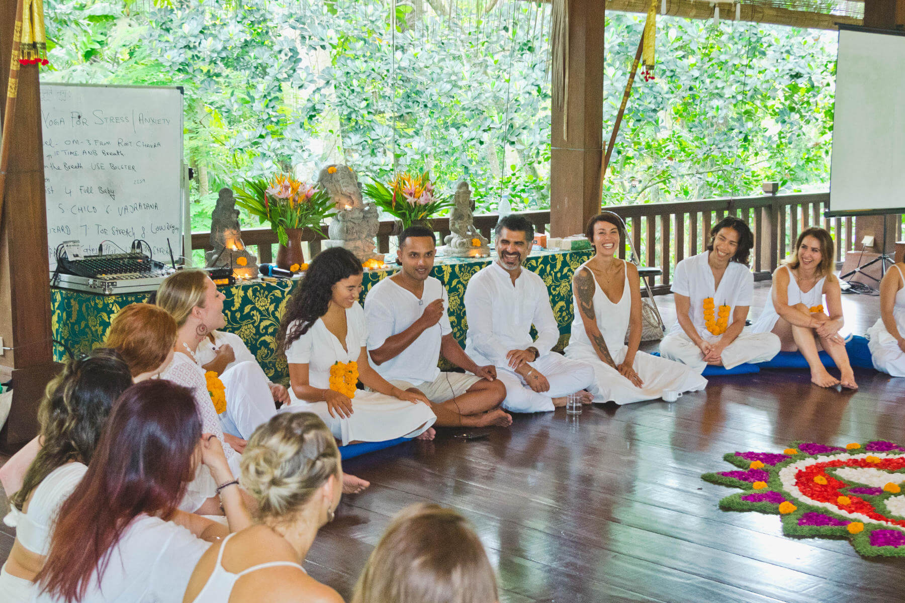 Group of people dressed in white, sitting in a circle at a yoga retreat at Namkhan Eco Farm in Luang Prabang, with flower decorations on the floor in a wooden-floored