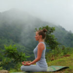 A woman practicing yoga in a seated pose on a mat outdoors, with misty mountains and lush greenery in the background of Namkhan, Luang Prabang.