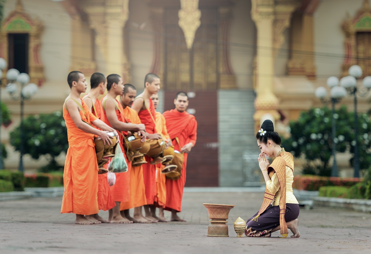 A woman in traditional attire kneeling and bowing before a group of monks in bright orange robes outside a temple in Luang Prabang.