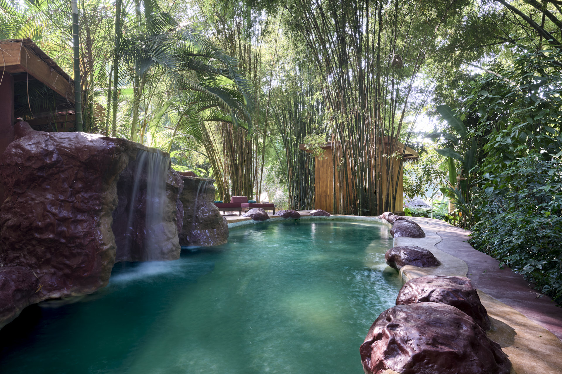 Outdoor hot spring pool surrounded by bamboo and tropical plants, with artificial waterfall and rock features, near a wooden building in Luang Prabang.