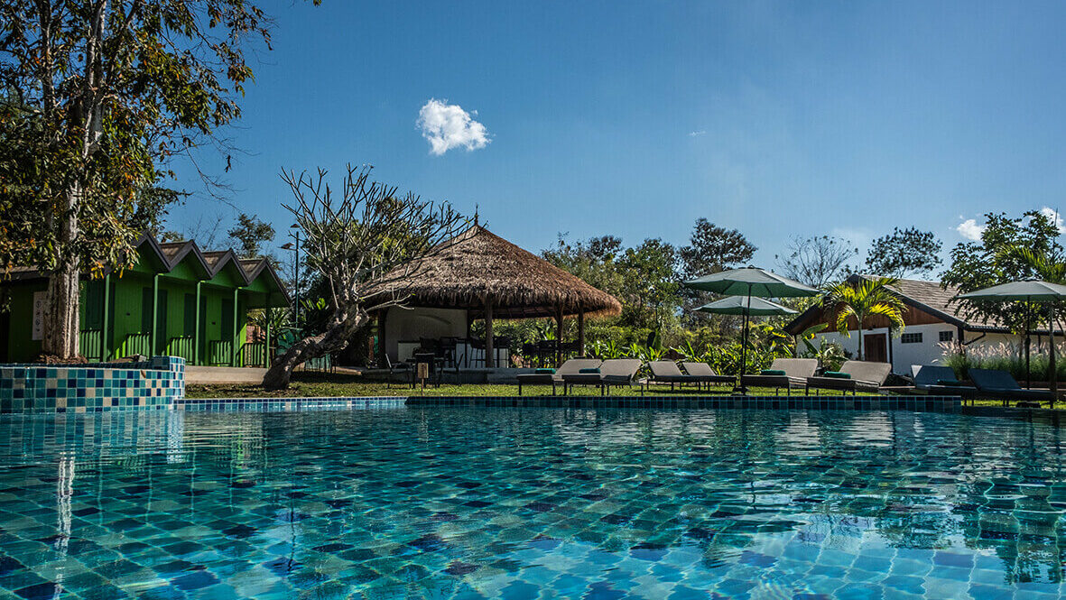 Outdoor tropical resort pool with blue tiles, lounge chairs, thatched roof cabana, and lush greenery under a clear sky in Luang Prabang.