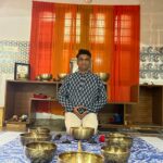 Hatha yoga teacher Vinayaka Honnavar training sitting cross-legged, surrounded by Tibetan singing bowls in a room with red curtains and decorative tiles.