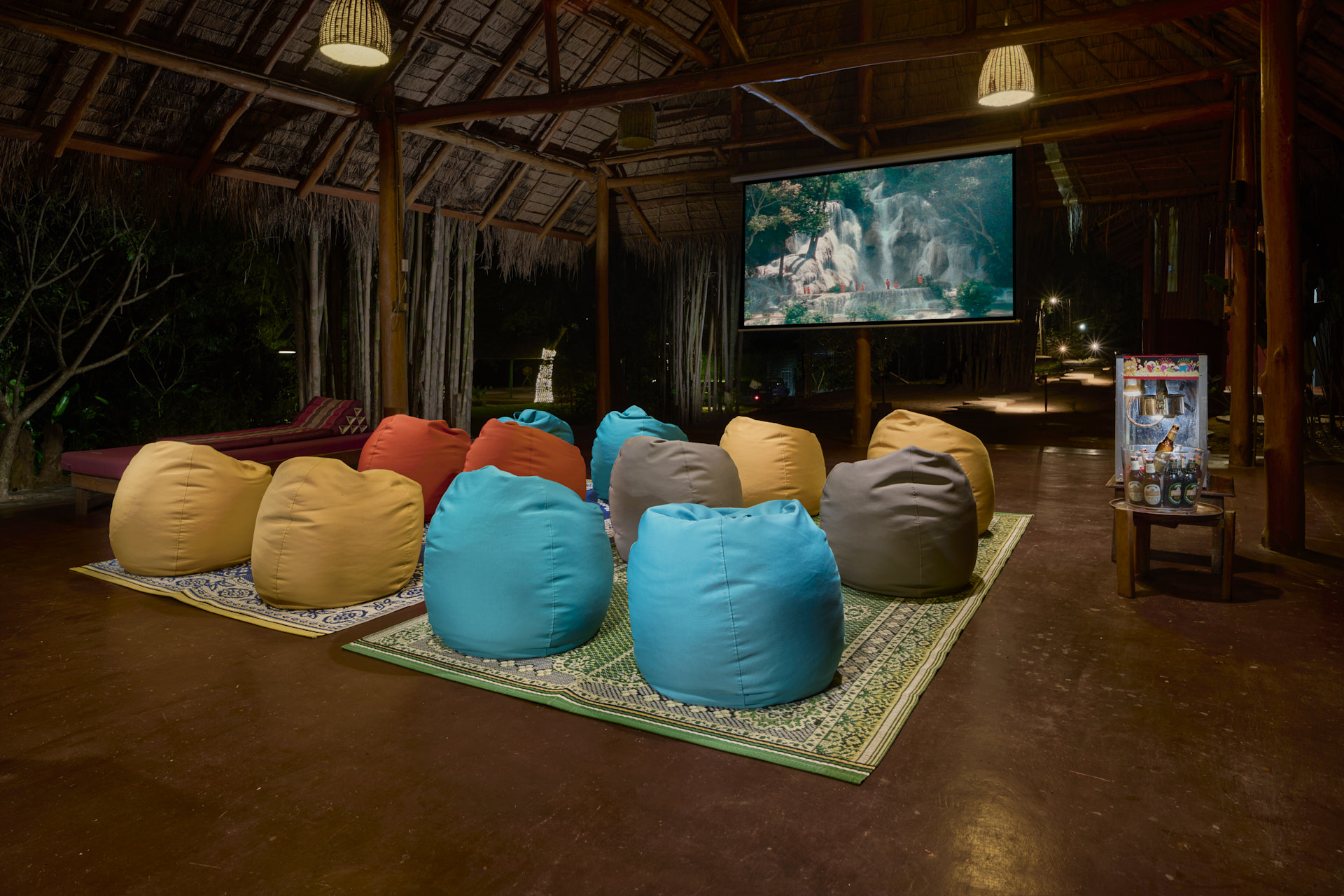 Colorful bean bags on a patterned rug in a dimly lit outdoor cinema at Namkhan Eco Farm, Luang Prabang, with a large screen displaying a movie, surrounded by lush