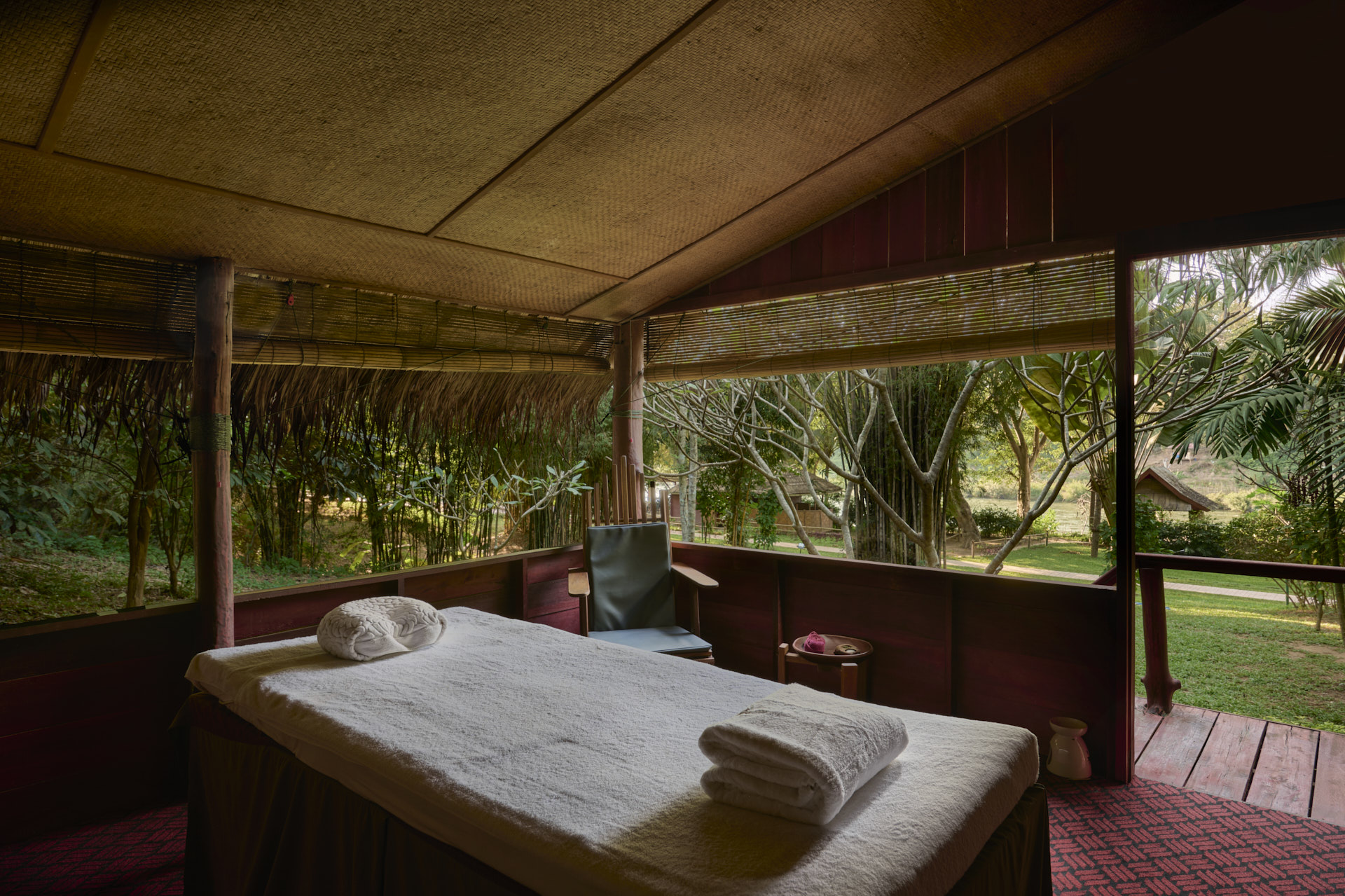Interior of a tranquil spa room with a massage table, folded towels, and a lush Namkhan garden view through bamboo blinds in Luang Prabang.