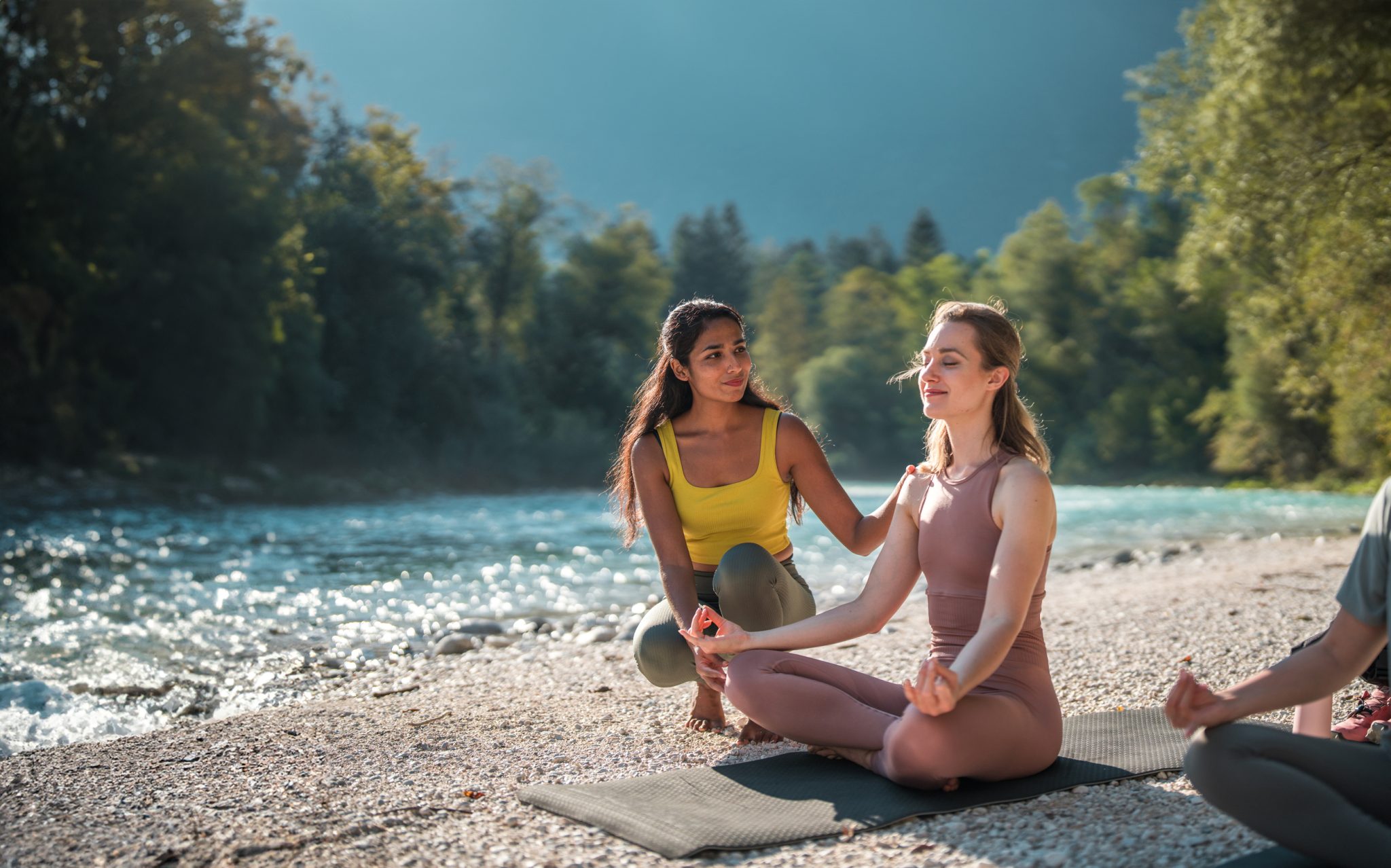 Two women practicing Ashtanga yoga on mats beside a river, surrounded by forested mountains, one in a yellow top and the other in a white bodysuit.