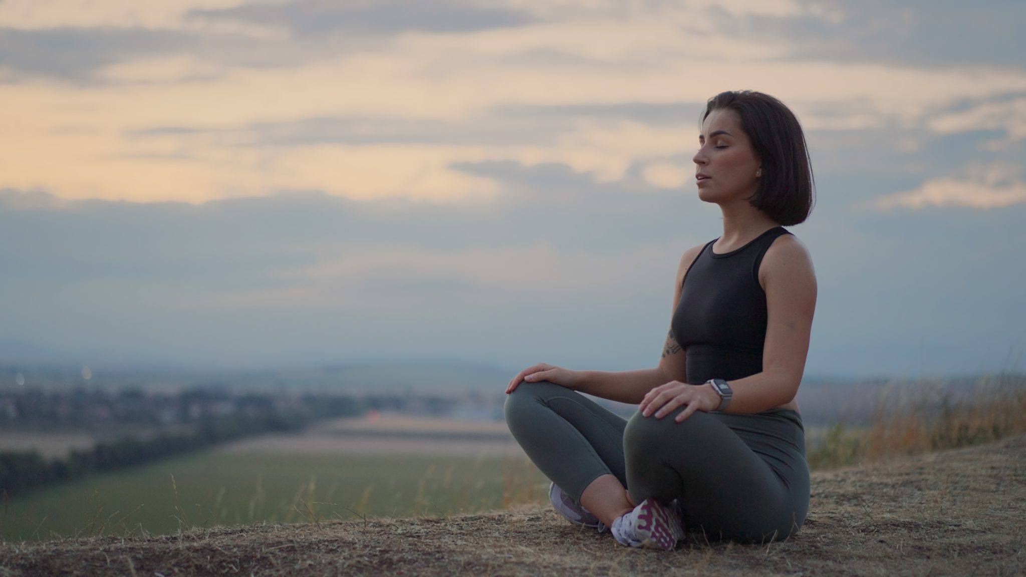 A woman participating in Ashtanga teacher training is meditating outdoors at dusk, sitting cross-legged on a hill with a serene expression and a soft-focus landscape in the background.